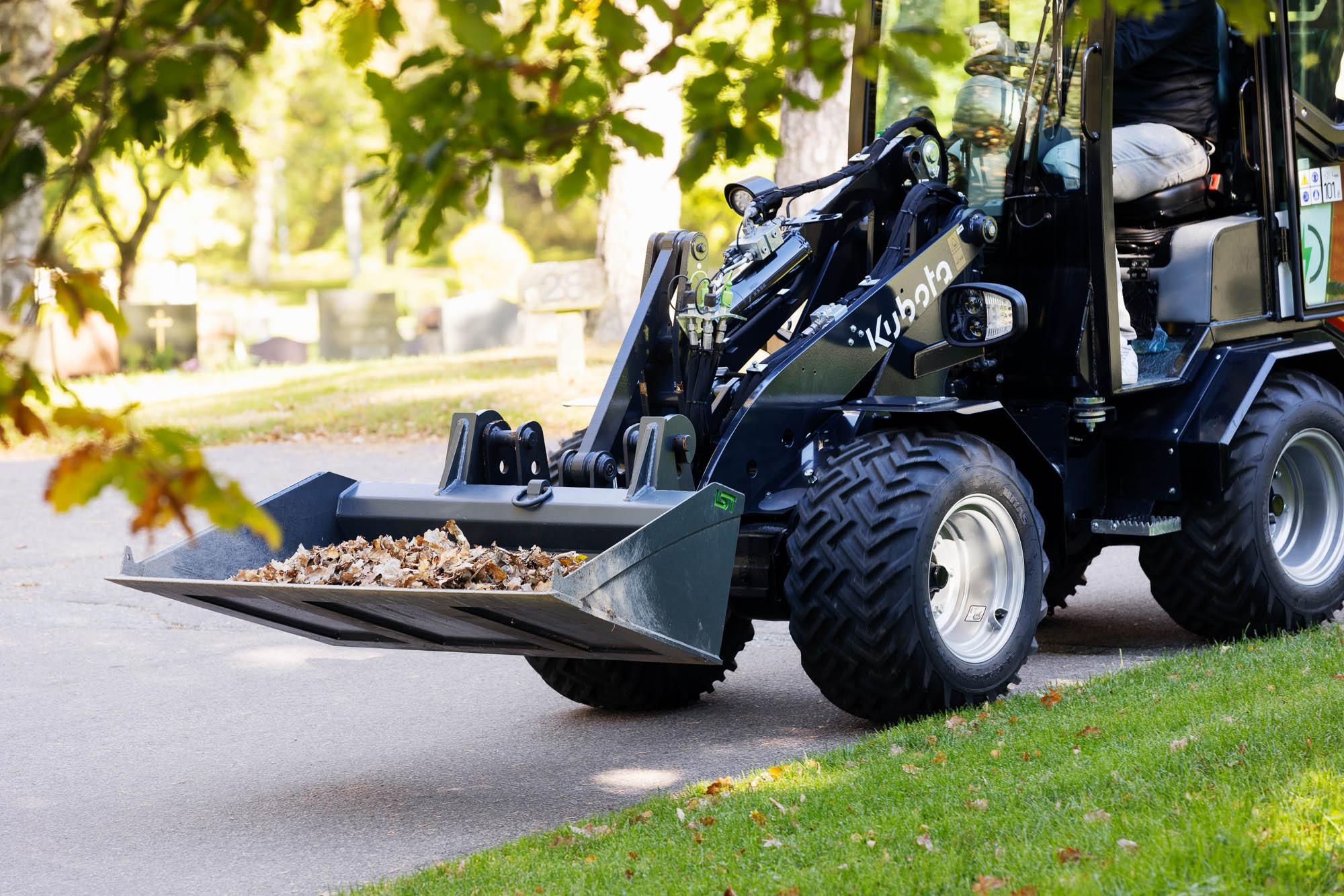 Kubota RT280-2e elektrisk hjullastare som transporterar löv på en kyrkogård, idealisk för tyst och avgasfri drift i känsliga miljöer.